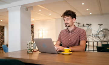 Man typing on a laptop. A yellow espresso cup and saucer sits on a table to his left.